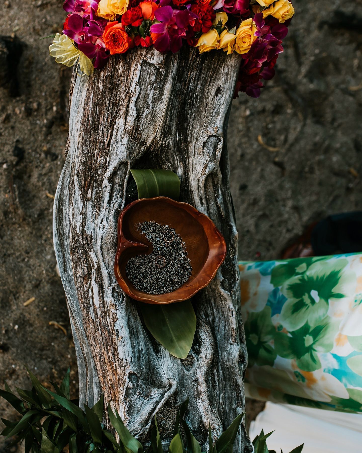 Rustic wooden stump with a small clay bowl, green leaf, colorful flowers, and a floral cloth backdrop