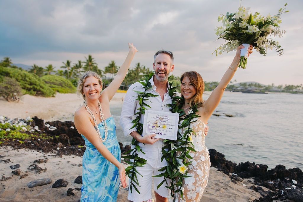 Three people celebrating by the water, holding bouquets and a certificate on a rocky beach.