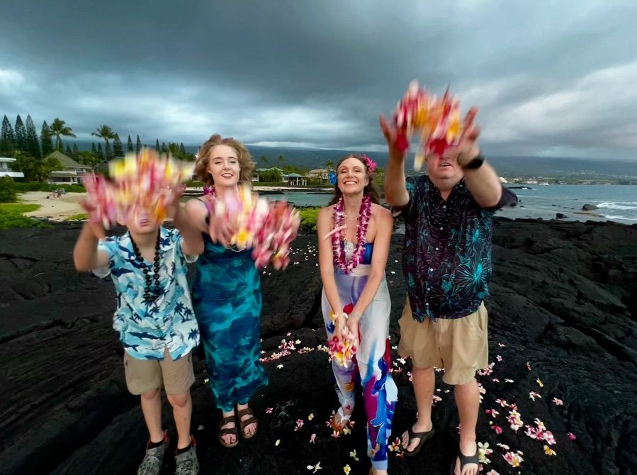 Four people on a rocky beach toss colorful petals at the camera under a cloudy sky.
