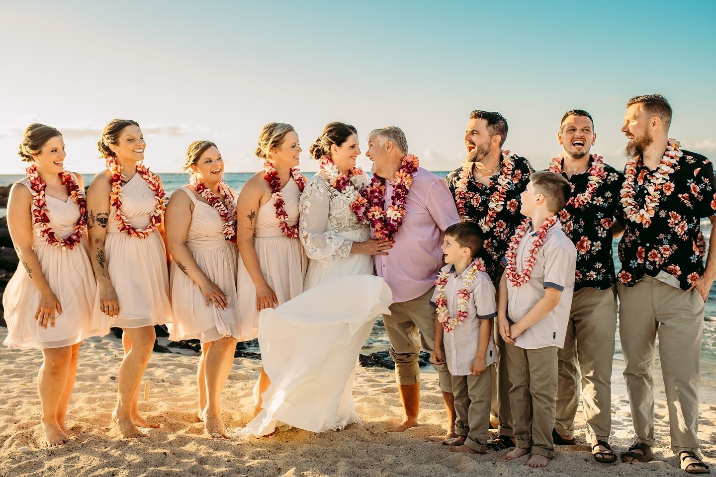 Wedding party on a beach. Bride and groom embrace, surrounded by bridesmaids and groomsmen wearing leis. Sunset light.