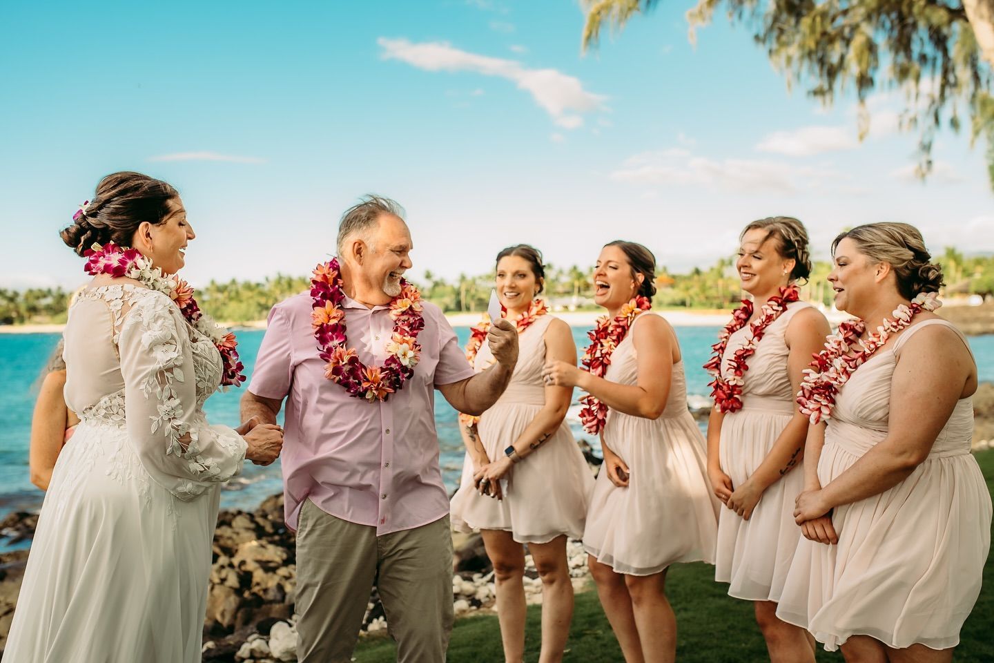 Wedding group smiles by ocean, bride in white dress, groom in pink shirt, bridesmaids in peach.