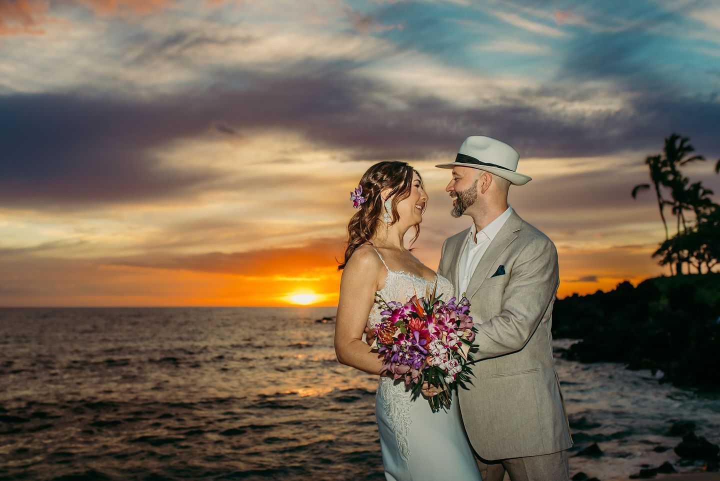 Bride and groom at sunset, embracing near ocean. Bride holds flowers; the groom wears a hat. Orange sky.