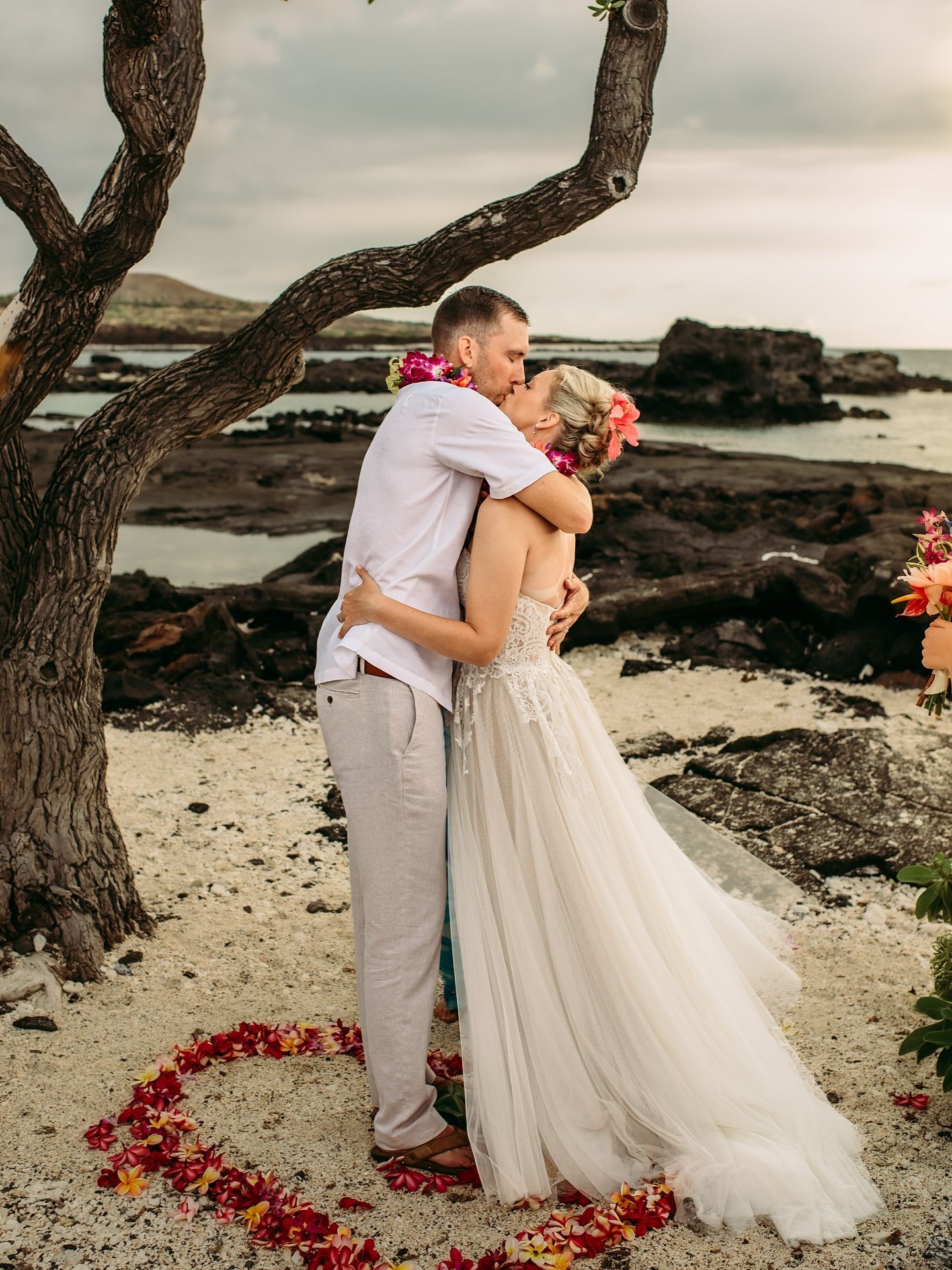 Couple kissing on a beach; she wears a wedding dress, he wears a shirt and pants. Flowers form a heart around them.