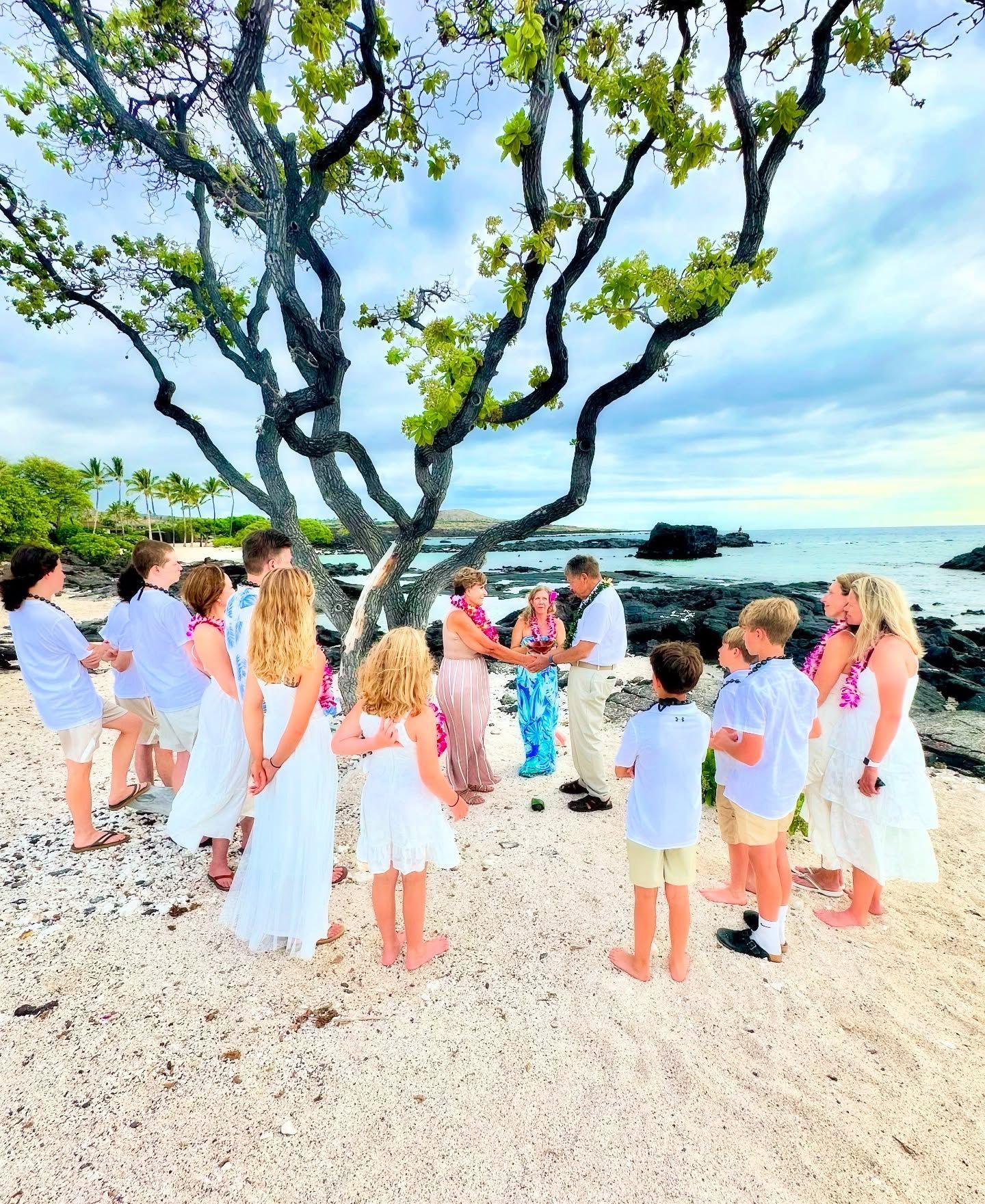 Group gathered on a beach under a large tree, facing the ocean in white clothing at a seaside ceremony.