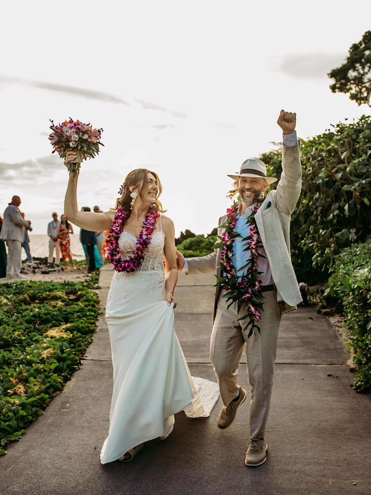 Newlyweds cheer, holding flowers and leis, walking on a path after a wedding ceremony.