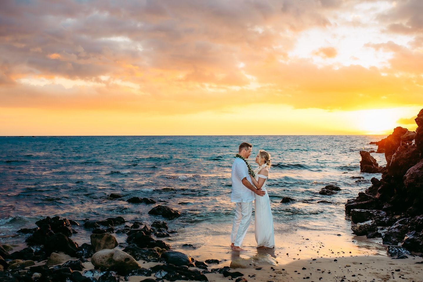 Couple embraces on beach at sunset; ocean, rocks, and colorful sky.