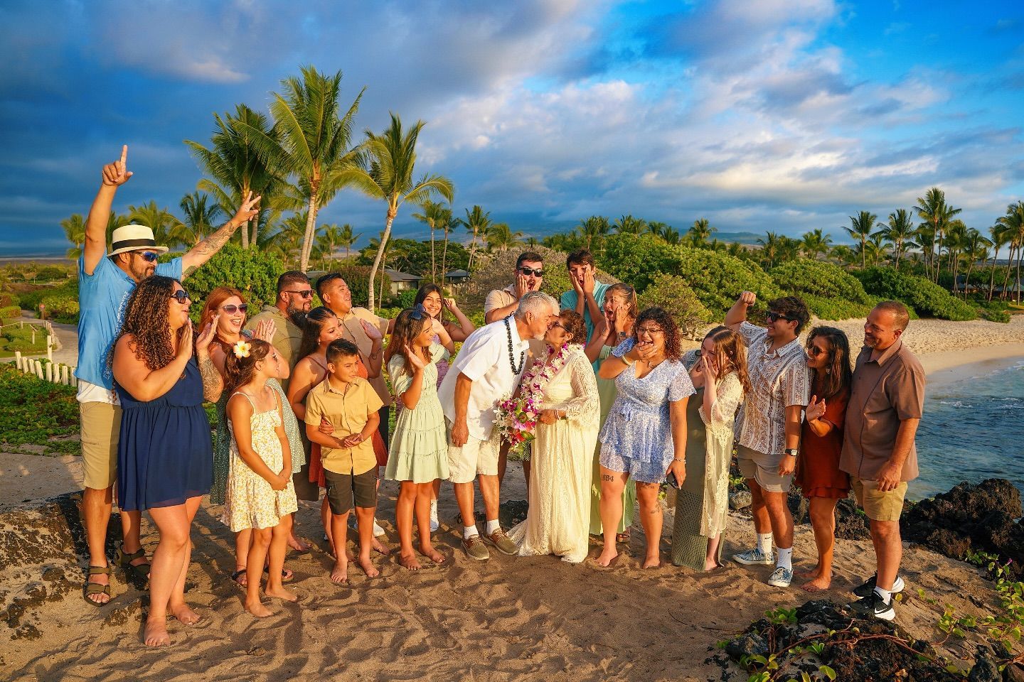 Group of people smiling on a beach; one man points up. Palm trees and blue sky background.