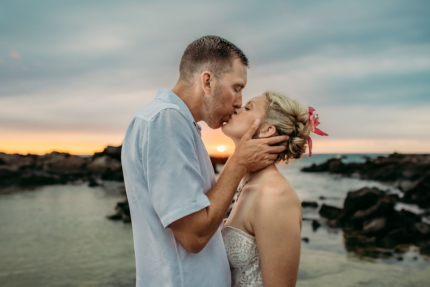 Couple kissing at sunset on a rocky shoreline. Woman wears a wedding dress, man in a light blue shirt.