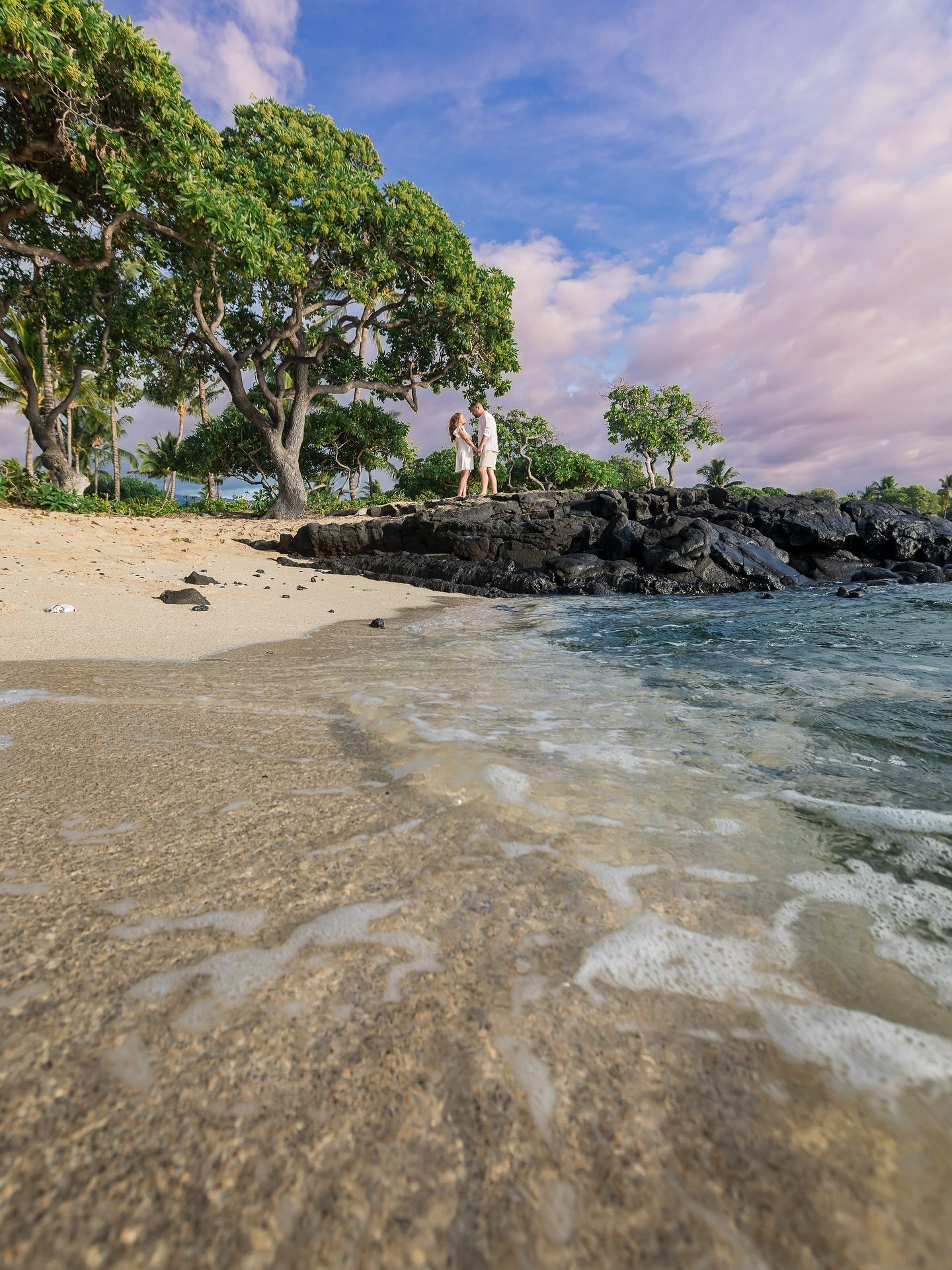 Sandy tropical beach with trees, black rocks, and gentle waves under a blue sky