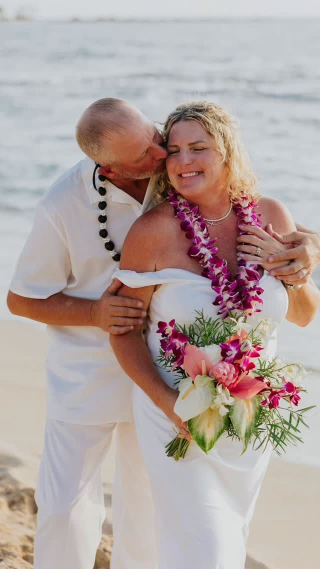 Couple in white on a beach, embracing with pink flower leis and bouquet by the ocean