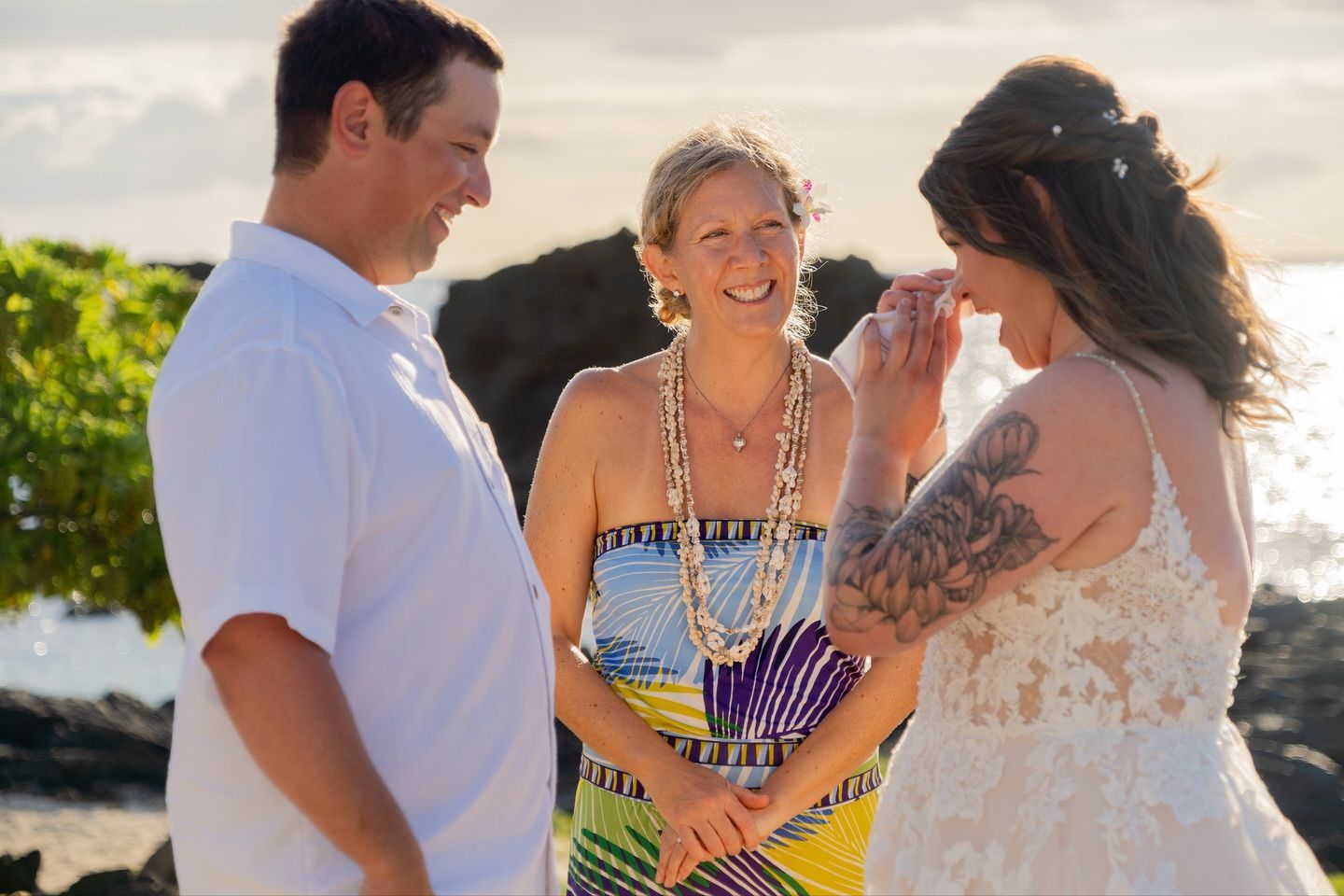 Couple at wedding ceremony; bride cries, officiant smiles, beach setting.