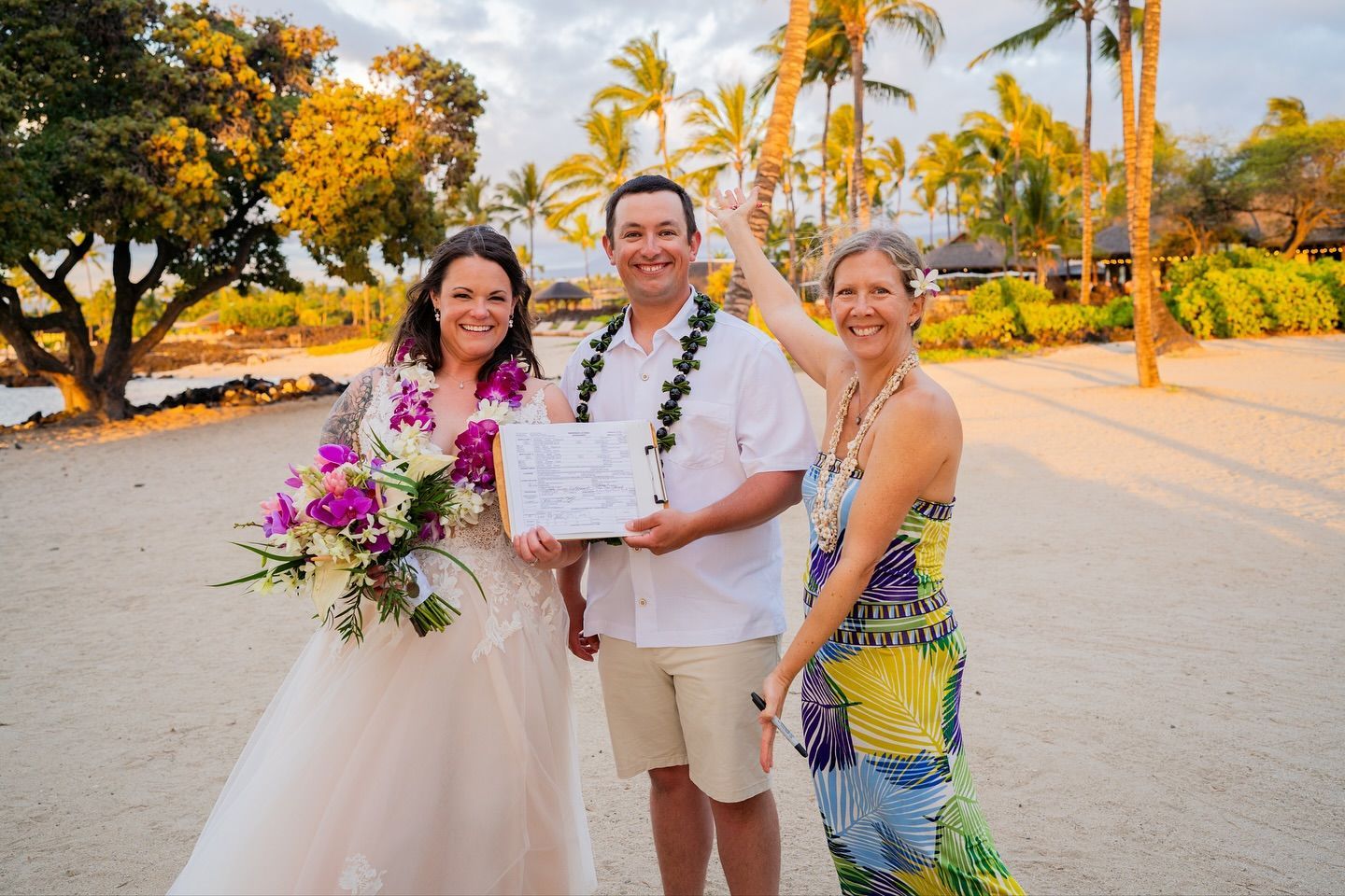Bride and groom hold marriage certificate on a beach with officiant. Bride holds bouquet, groom wears lei.