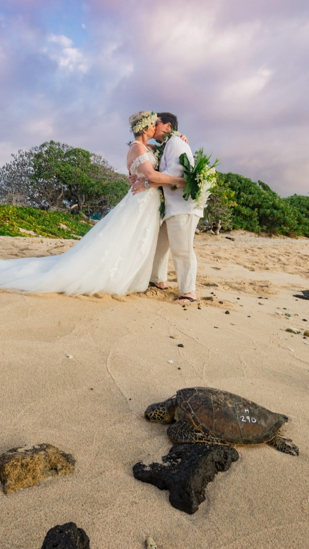 Couple in wedding attire kissing on a sandy beach with palm trees and cloudy sky