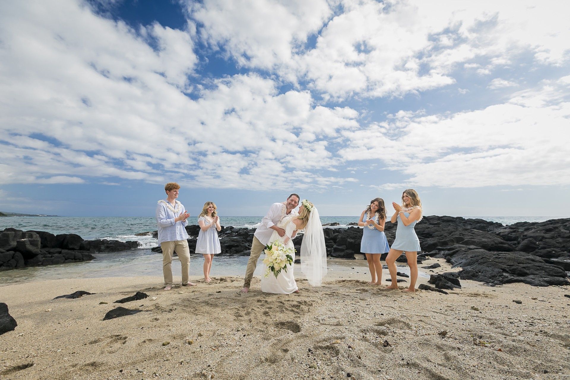 Wedding party posing on a rocky beach beneath a cloudy sky, with the bride and groom centered.