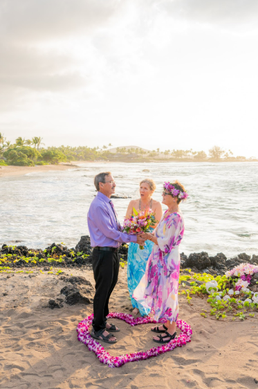 Couple embraces on a beach, woman in wedding dress and floral crown, man in white shirt and tan pants.
