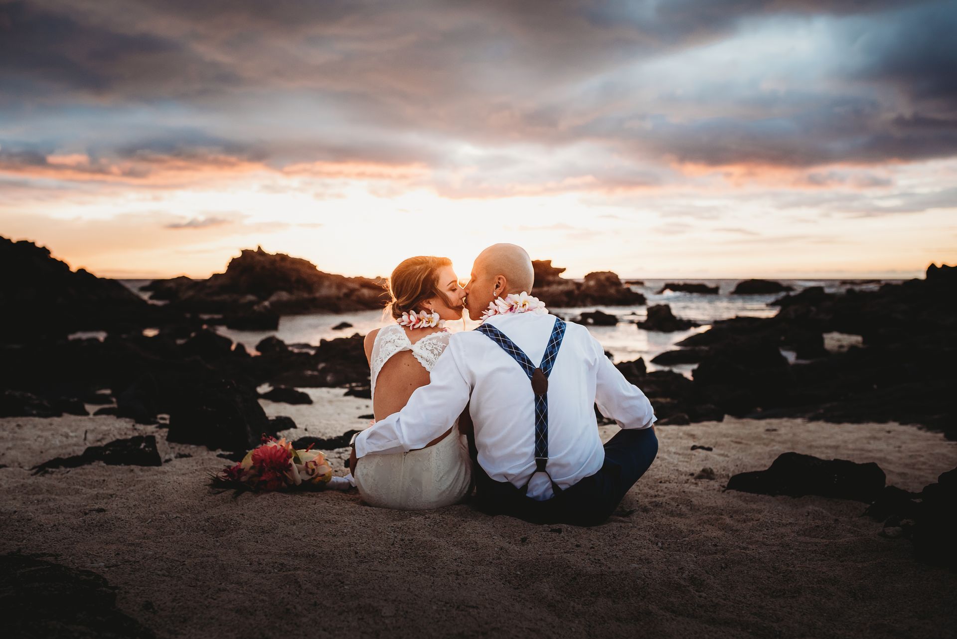 Simple Kona Beach Weddings couple, sitting on the beach, facing the sunset, lava and ocean in the background, kissing. They are both wearing white, and he has on black suspenders.
