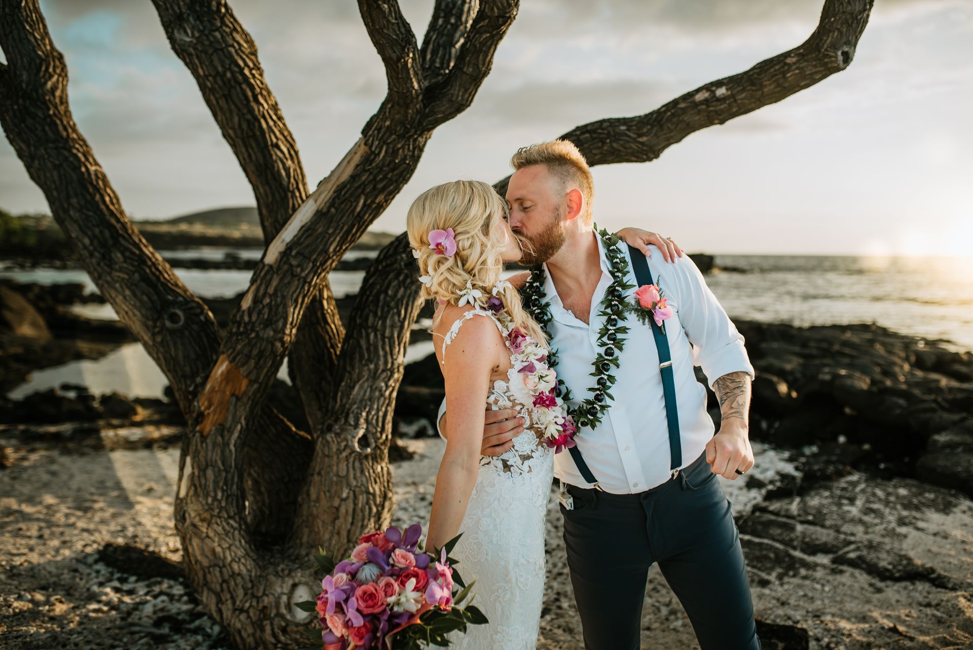 A Simple Kona Beach Wedding couple in wedding attire kissing by a coastal tree, with bouquet and ocean at sunset