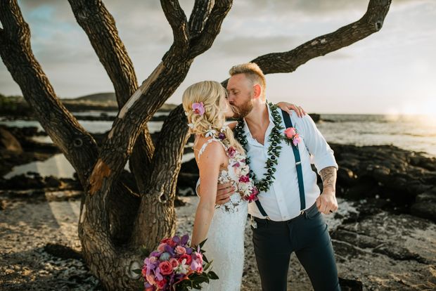 A Simple Kona Beach Wedding couple in wedding attire kissing by a coastal tree, with bouquet and ocean at sunset