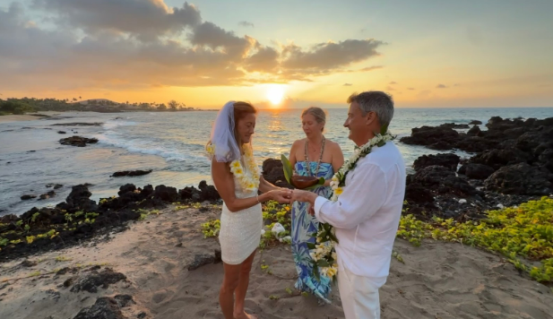 Couple wearing leis on beach at sunset. Woman smiles, holds flowers; man in hat smiles.
