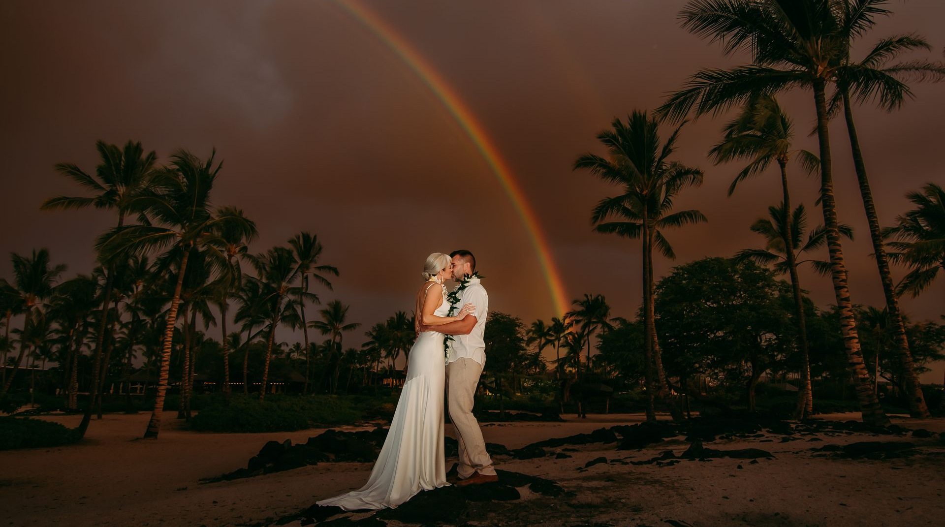 A Simple Kona Beach Weddings couple embracing on a beach at sunset with palm trees and a rainbow overhead