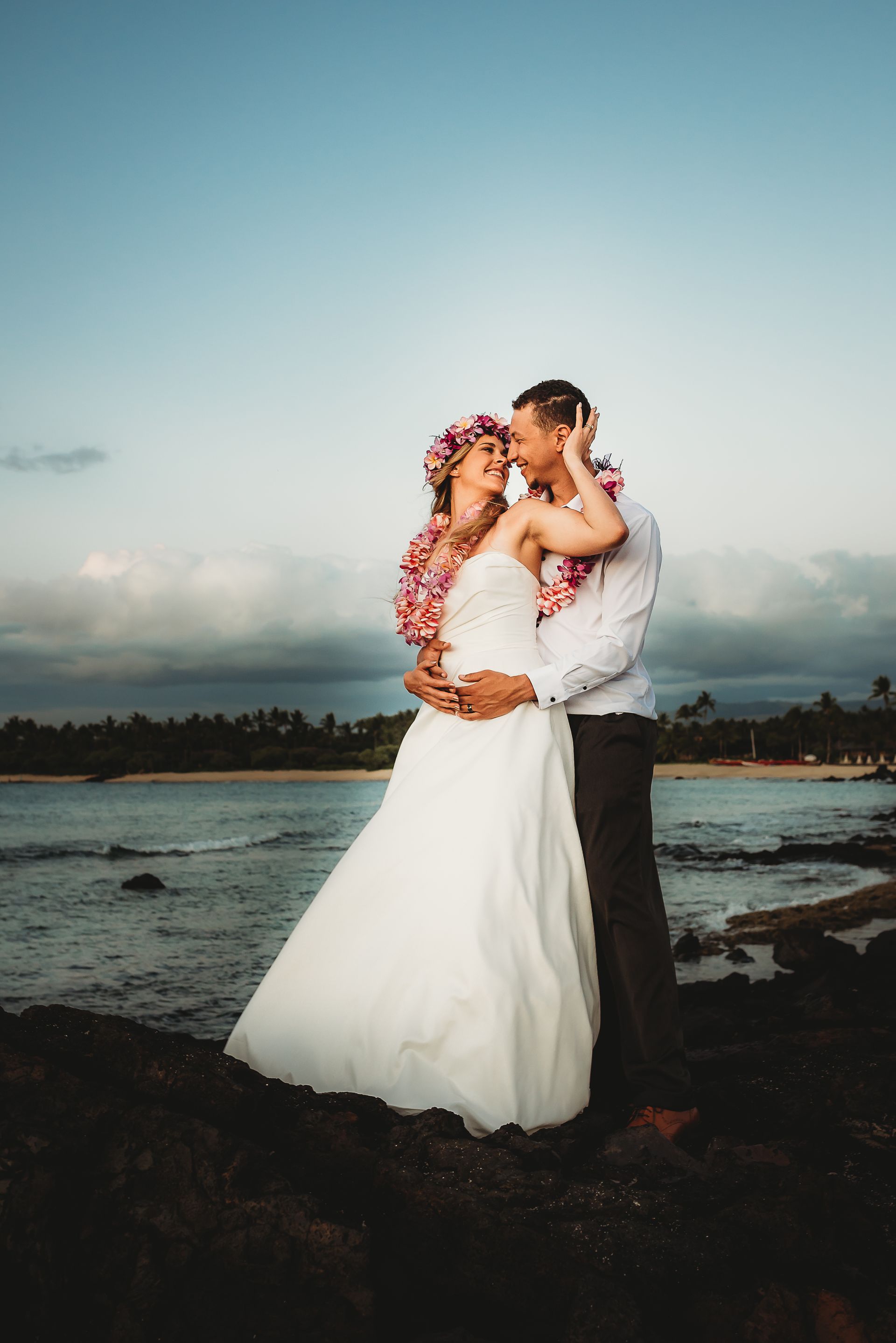 Couple embracing on a rocky beach at sunset, bride in white gown and flower crown, groom in white shirt.