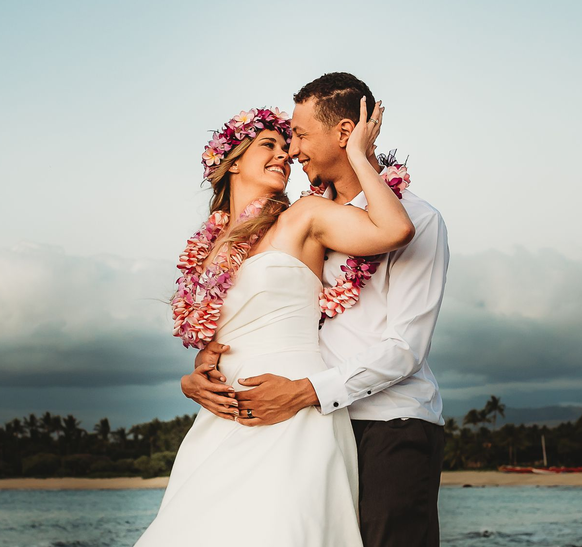 Couple embracing on a beach at sunset, wearing white with pink floral leis, smiling at each other