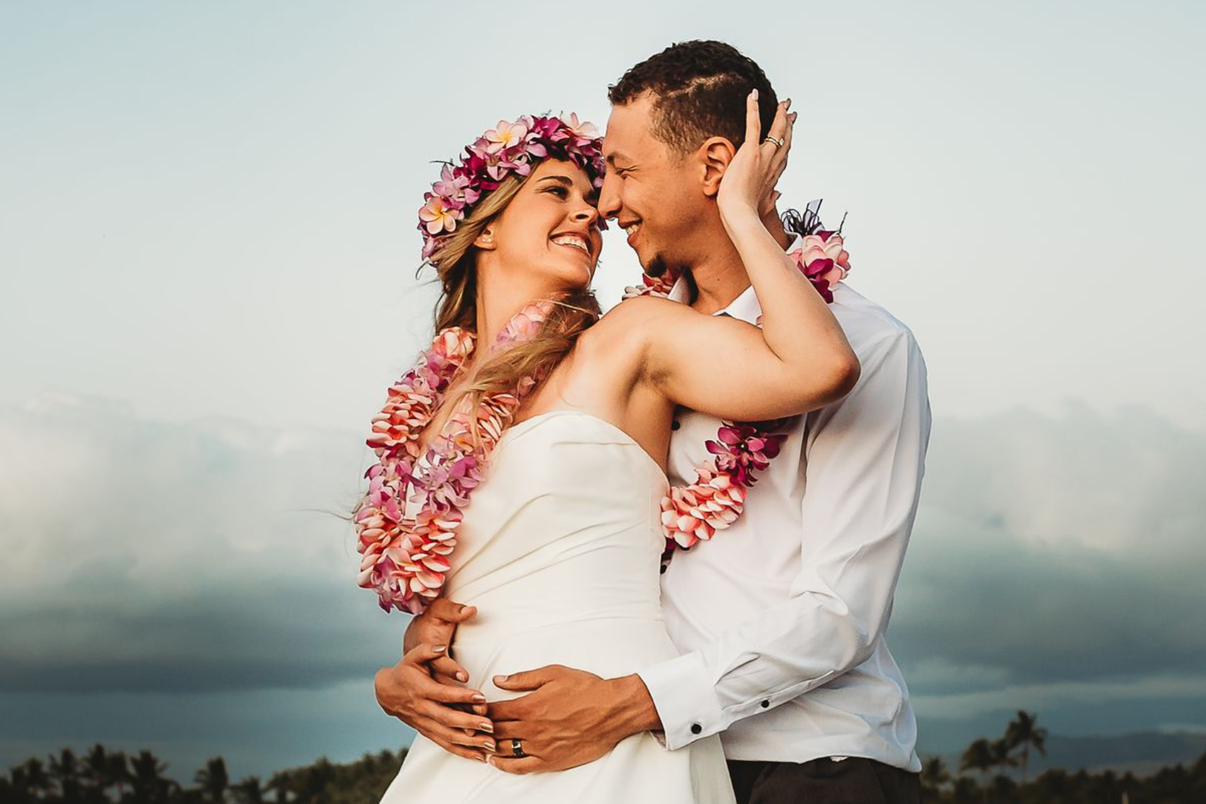 A Simple Kona Beach Wedding couple, on a beach in Kailua-Kona on the Big Island of Hawaii, wearing tropical flowers. The groom is standing behind the bride with his arms around her waist, and she is turned back, cupping his head with her hand. They are both smiling.