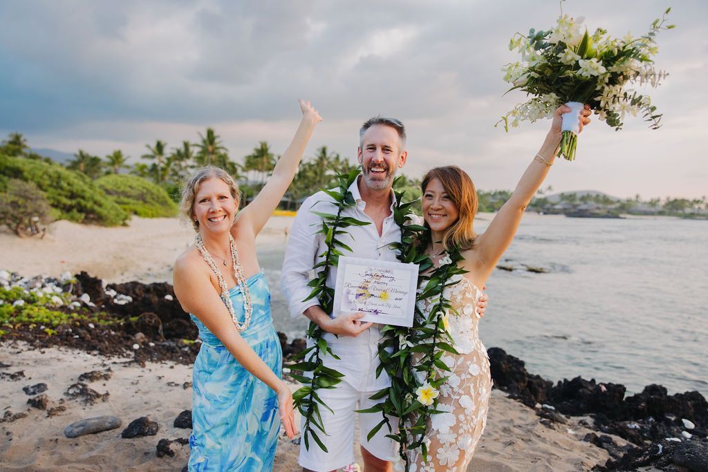 Three people celebrating on a beach after A Simple Kona Beach Wedding, smiling and holding up a bouquet and certificate.