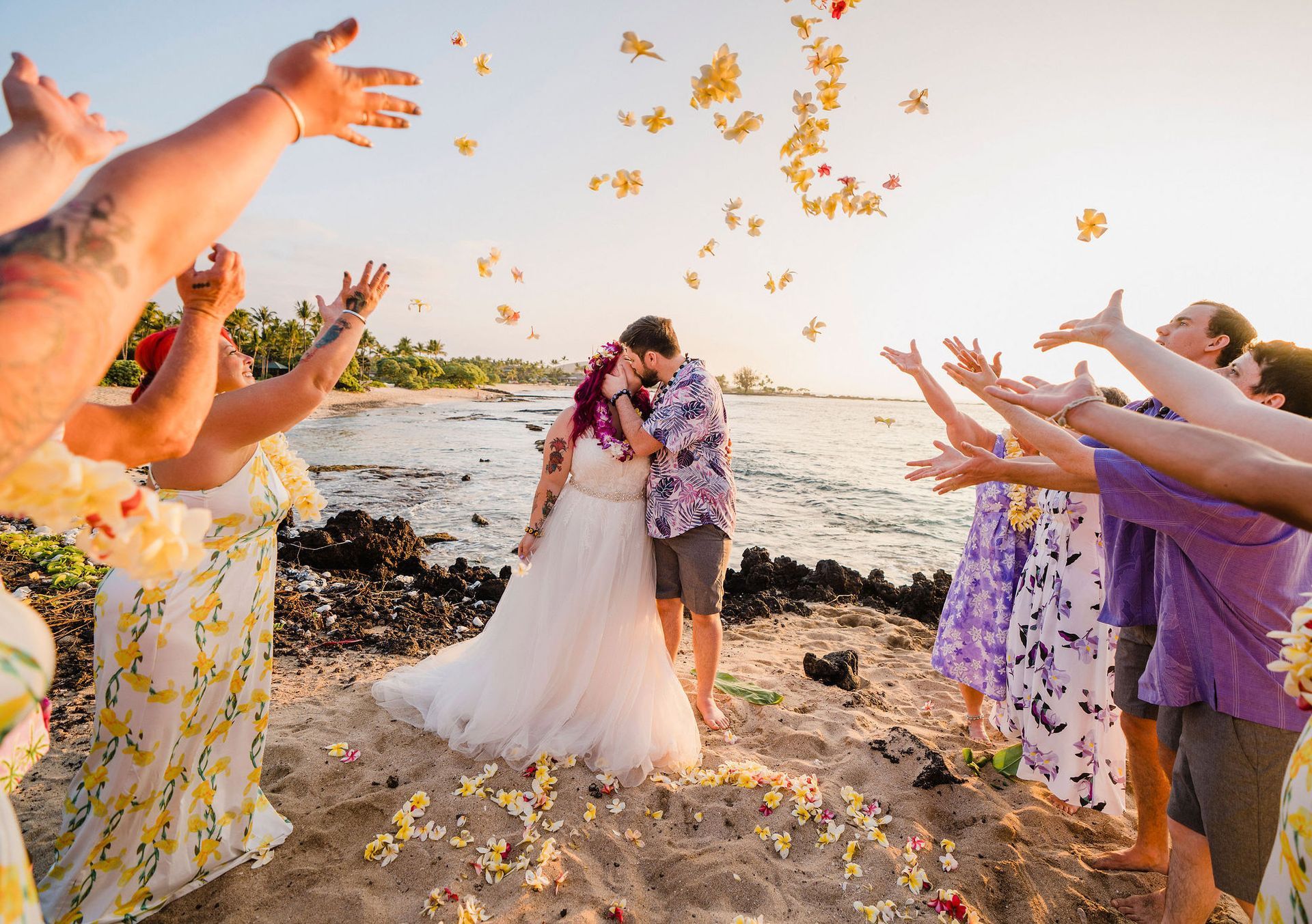 Bride and groom kiss on a beach as guests toss flower petals around them after a Simple Kona Beach Wedding ceremony.
