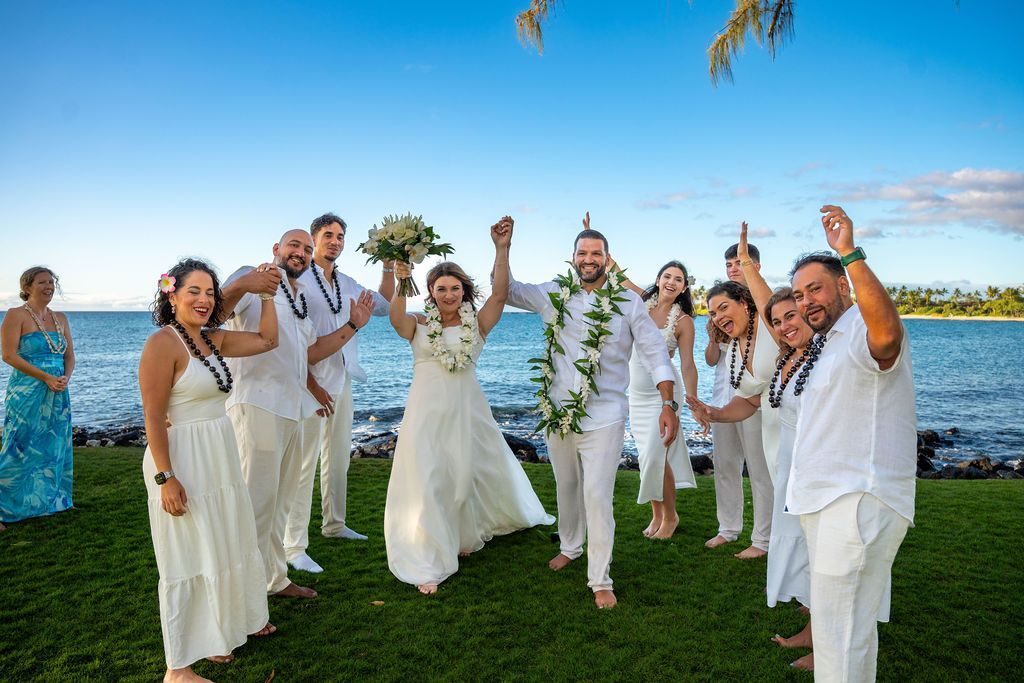 Wedding party in white celebrating on a grassy beach with the ocean behind them.