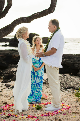 Couple embraces on a beach, woman in wedding dress and floral crown, man in white shirt and tan pants.