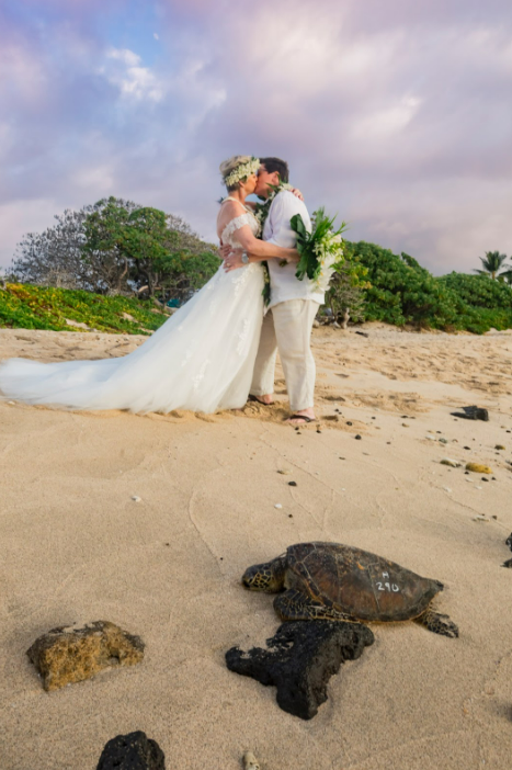 Couple wearing leis on beach at sunset. Woman smiles, holds flowers; man in hat smiles.