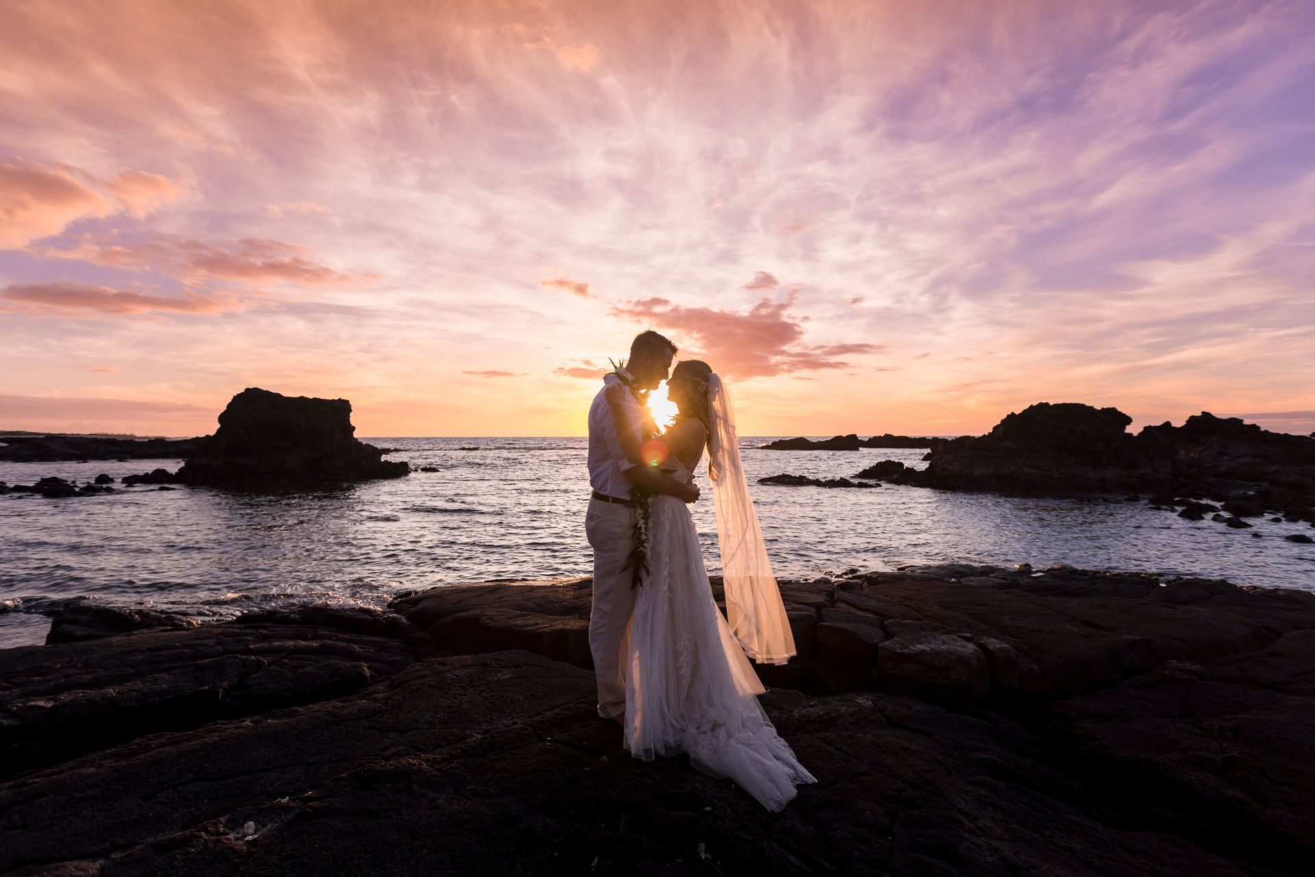 A Simple Kona Beach Wedding couple embracing in wedding attire at sunset by the rocky seashore
