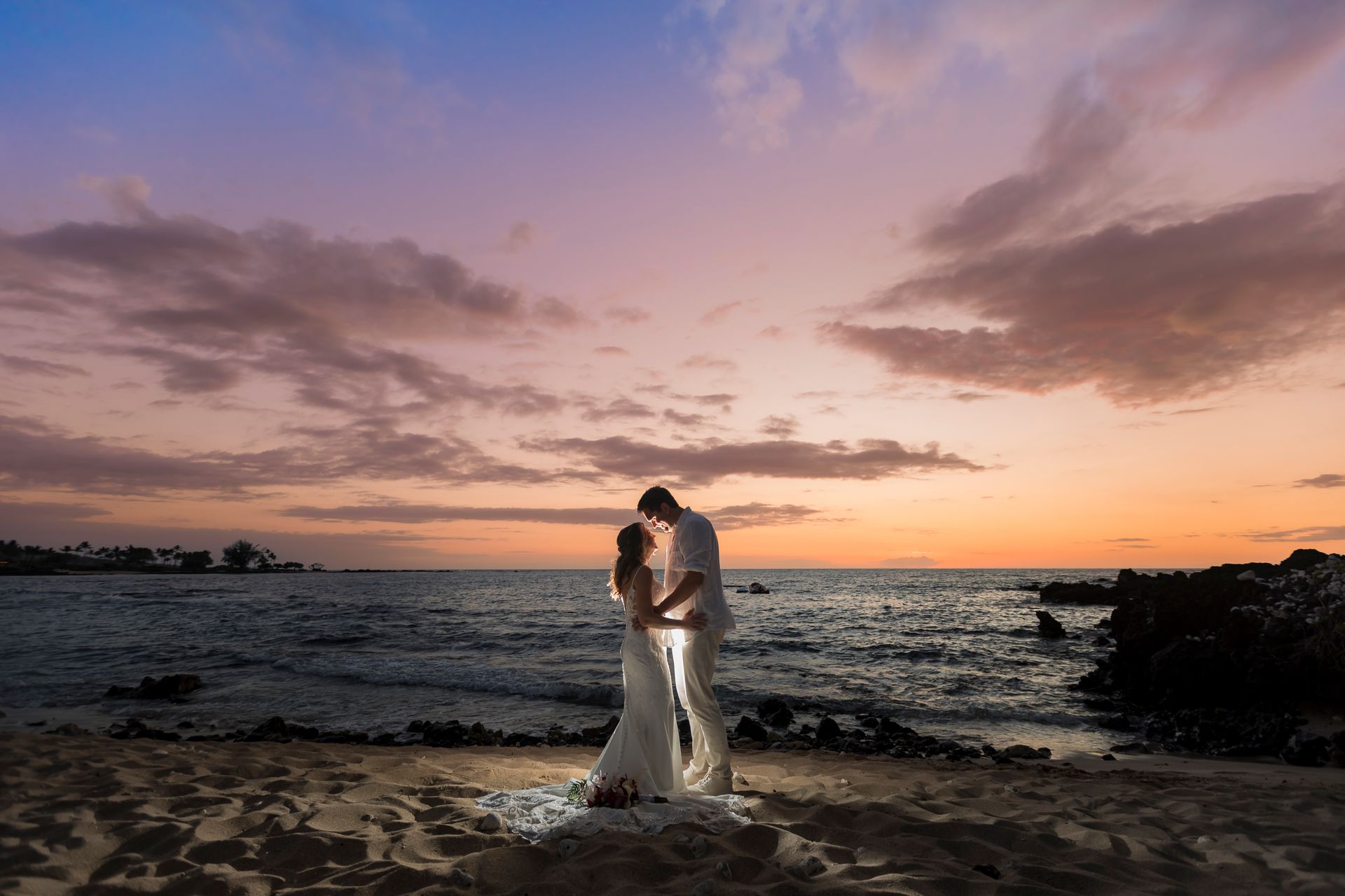 A Simple Kona Beach Weddings couple, standing on a sandy Big Island beach, with a colorful sunset in the background, facing one another. The couple is wearing white, and they are lit up in the middle.