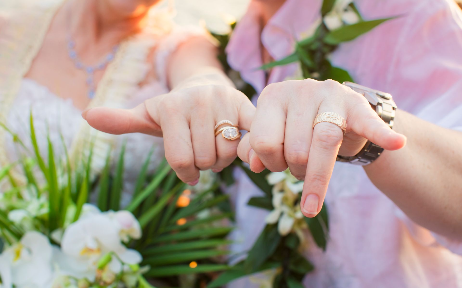 Hands with rings making a heart shape above greenery and pink flowers