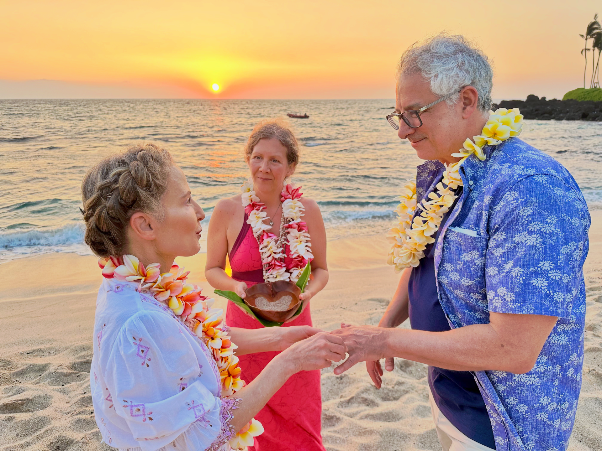 Couple embraces on a beach, woman in wedding dress and floral crown, man in white shirt and tan pants.