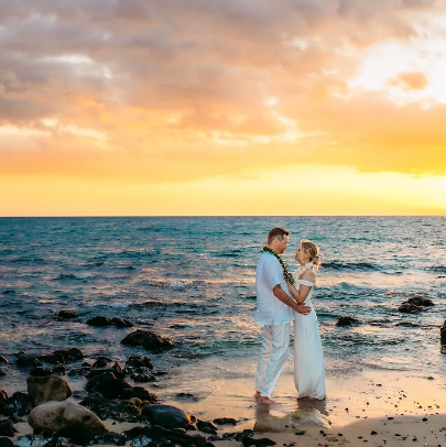 Couple barefoot on a sandy beach, surrounded by lava rocks, embracing at sunset.