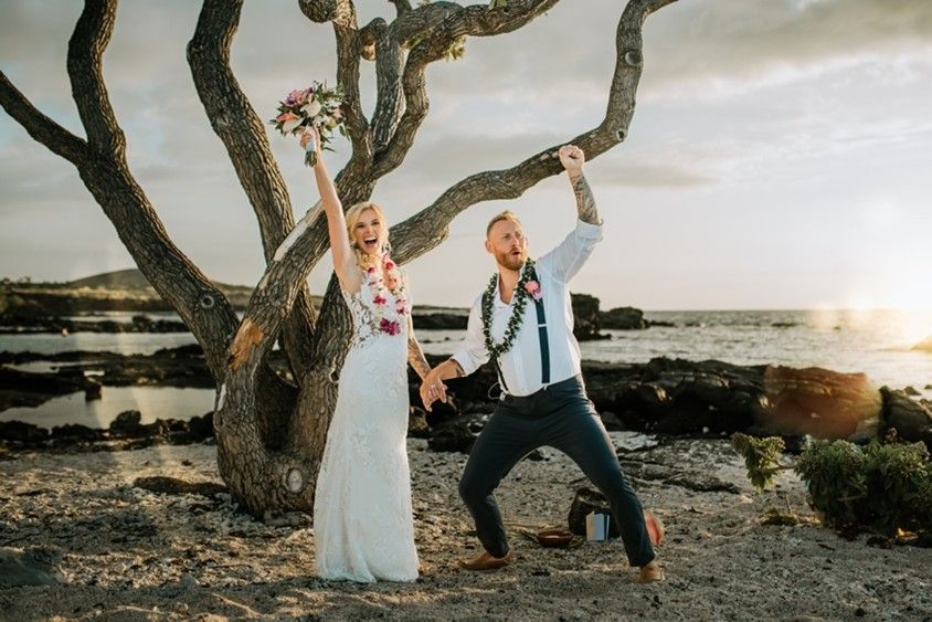 A couple raises their hands in celebration after their Hawaiian-style outdoor beach wedding ceremony