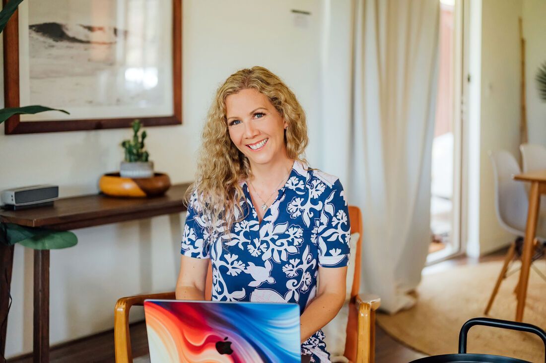 Becky Ringler of Simple Kona Beach Weddings smiles, seated with laptop in a room with wood table, plants, and natural light.