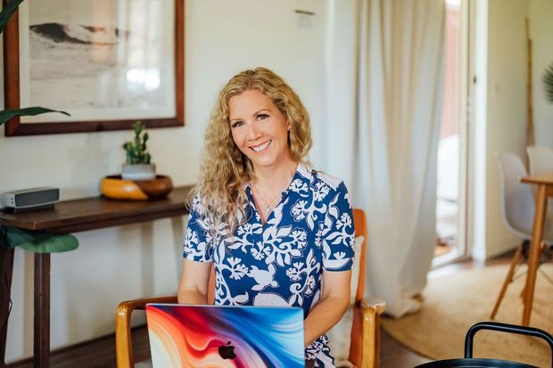 Becky Ringler of Simple Kona Beach Weddings smiles, seated with a laptop in a sunlit room in Kailua-Kona, HI.