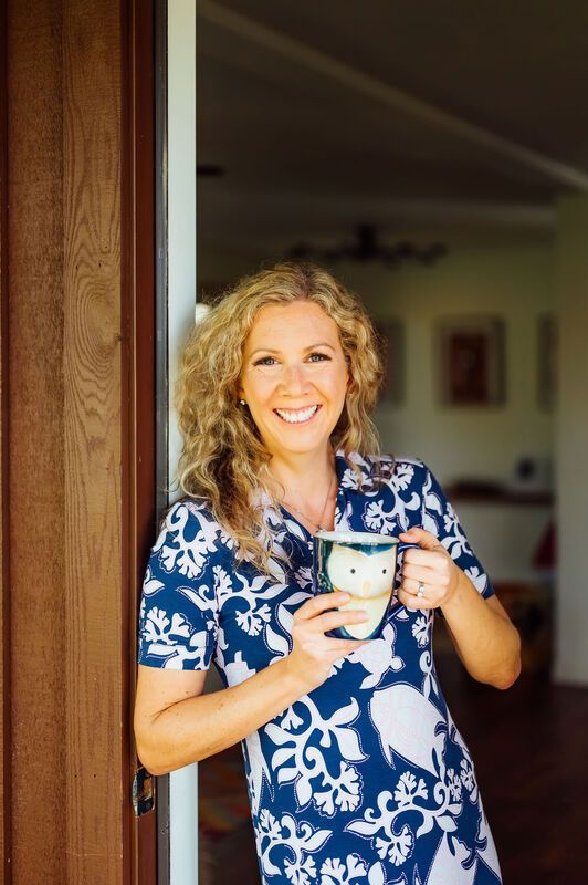 Woman smiles holding a mug, standing in a doorway; wearing a blue and white patterned shirt.