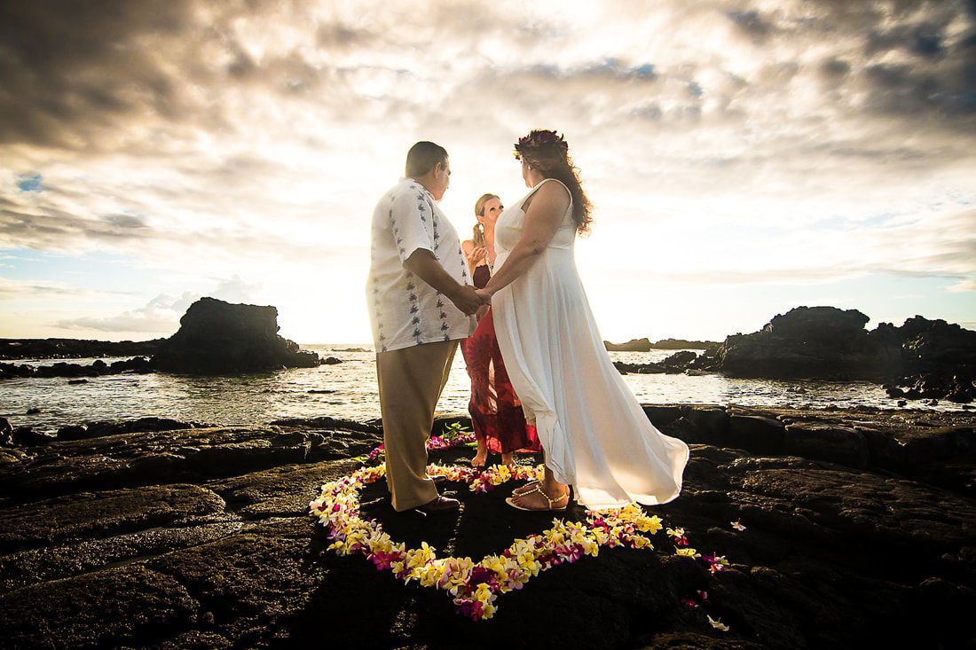 A Simple Kona Beach Wedding couple holding hands on a rocky shoreline at sunset, with bright floral arrangements in the foreground