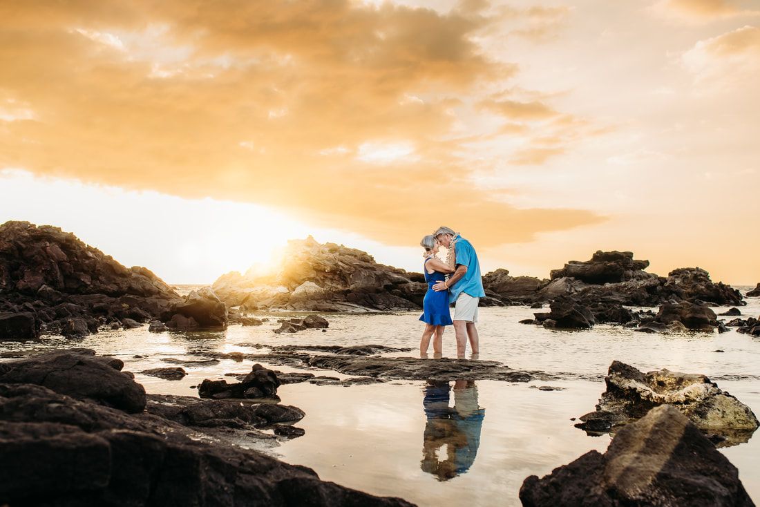 Couple embracing on a rocky beach at sunset, with warm orange sky and reflections in shallow water