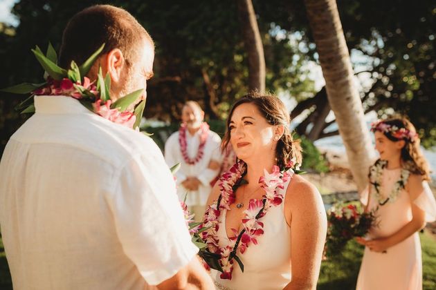 Simple Kona Beach Weddings Custom Vow Renewal. A couple exchanging vows during a Hawaiian vow renewal ceremony, wearing leis. Attendees in the background.