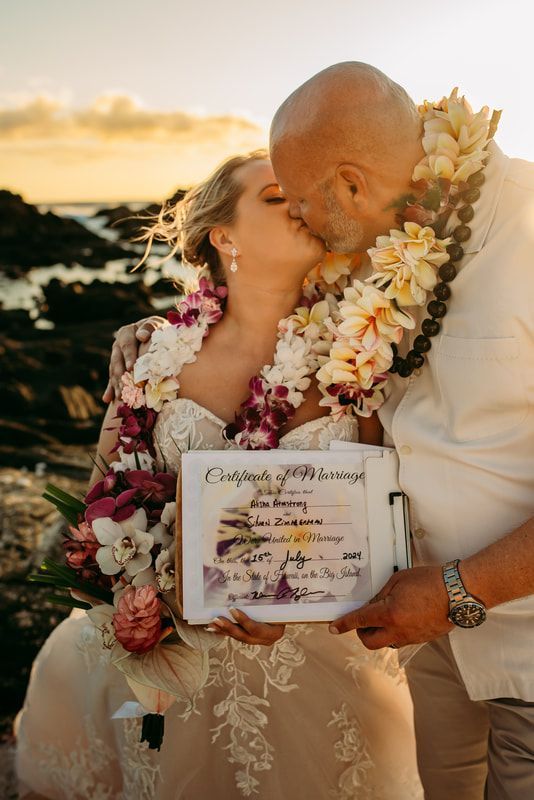 Bride and groom kissing, holding marriage certificate near the ocean at sunset; both wearing leis.