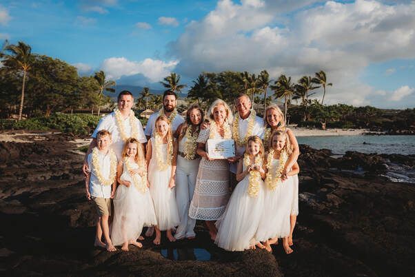Simple Kona Beach Weddings vow renewal couple, posing with their family on the lava on a Big Island beach with palm trees in the background. Sunset lighting and blue sky. All wearing white, donning yellow plumeria leis.