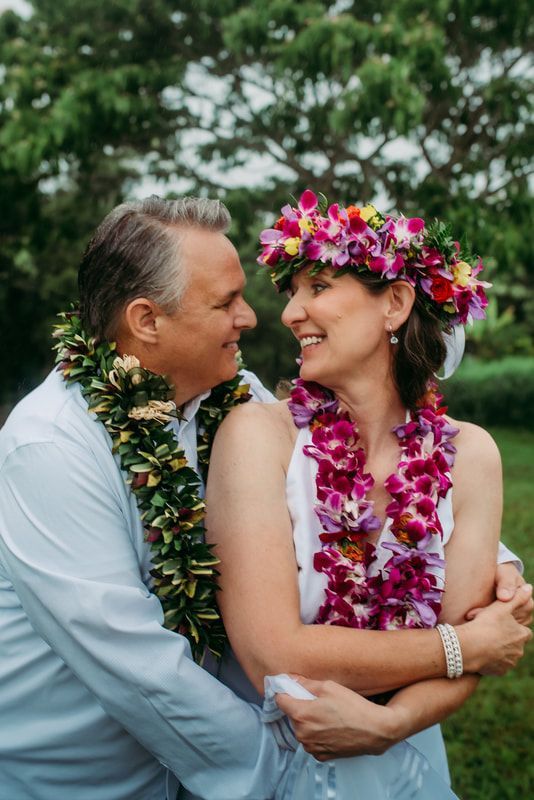 Couple embracing, smiling at each other, wearing floral leis. Outdoor, green backdrop.