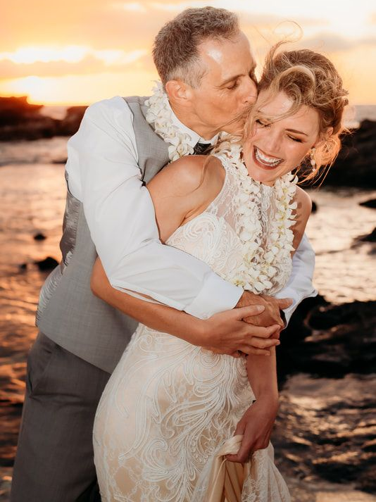 Couple embracing on a beach at sunset, with the woman in a lace dress smiling as the man kisses her cheek