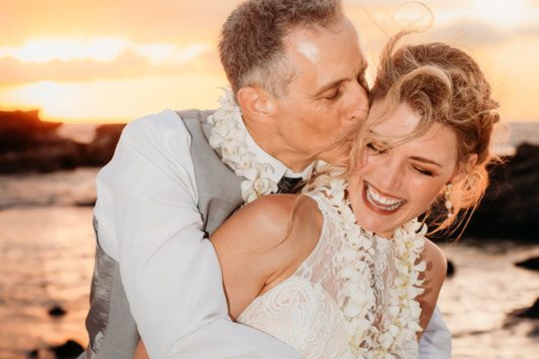A Simple Kona Beach Wedding couple, at sunset on the beach on the Kona Coast on the Big Island of Hawaii, the groom kissing the bride on the cheek while the bride smiles and laughs.