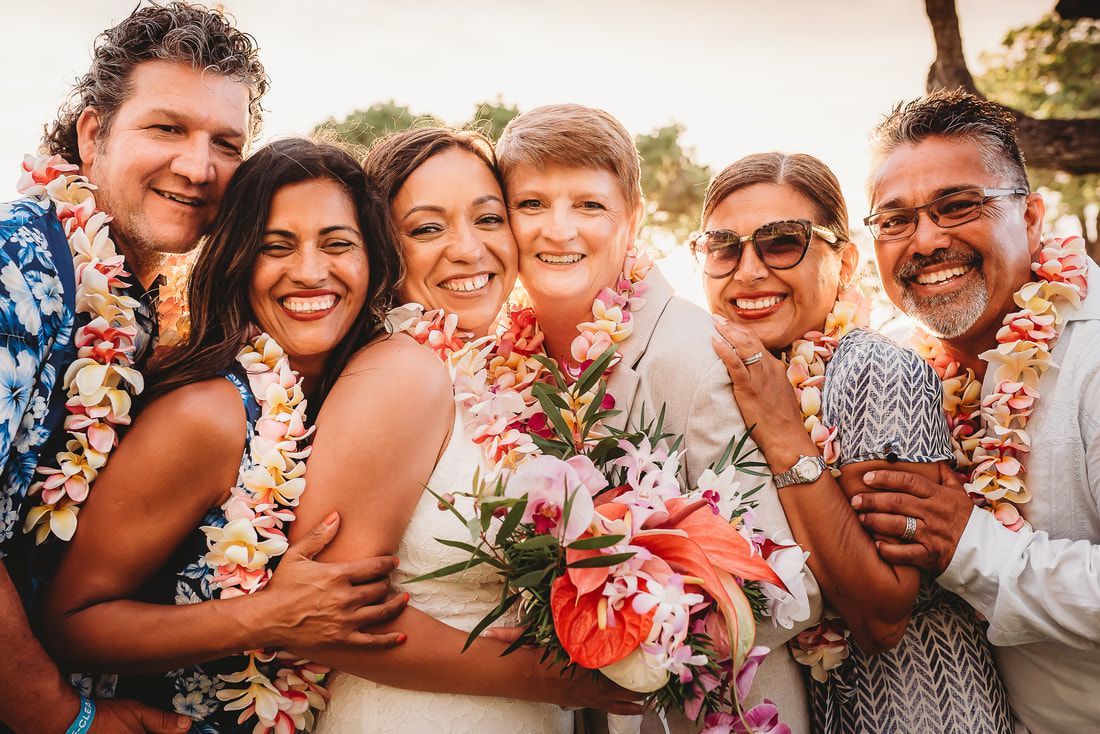 Smiling group wearing flower leis pose together outdoors at sunset, holding a bouquet of pink and white flowers